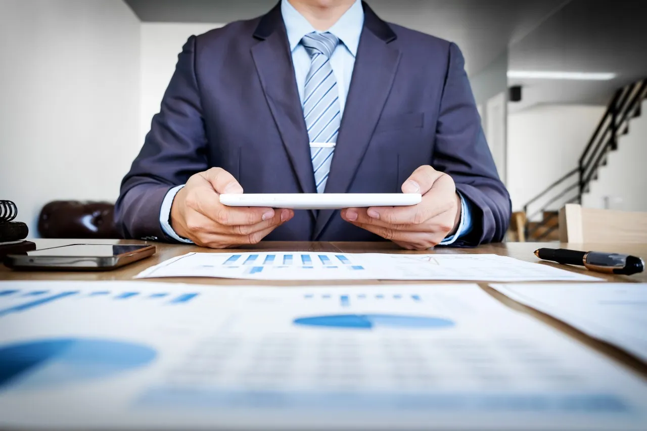 A businessman in a suit holds a tablet over financial charts on a desk.