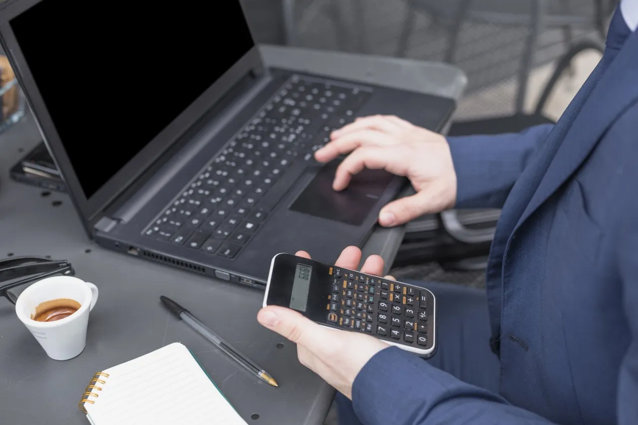 From a high angle, a person in a blue suit uses a laptop with a blank screen and holds a calculator in their other hand. A cup of coffee, glasses, a pen, and a notebook are also on the grey outdoor table.