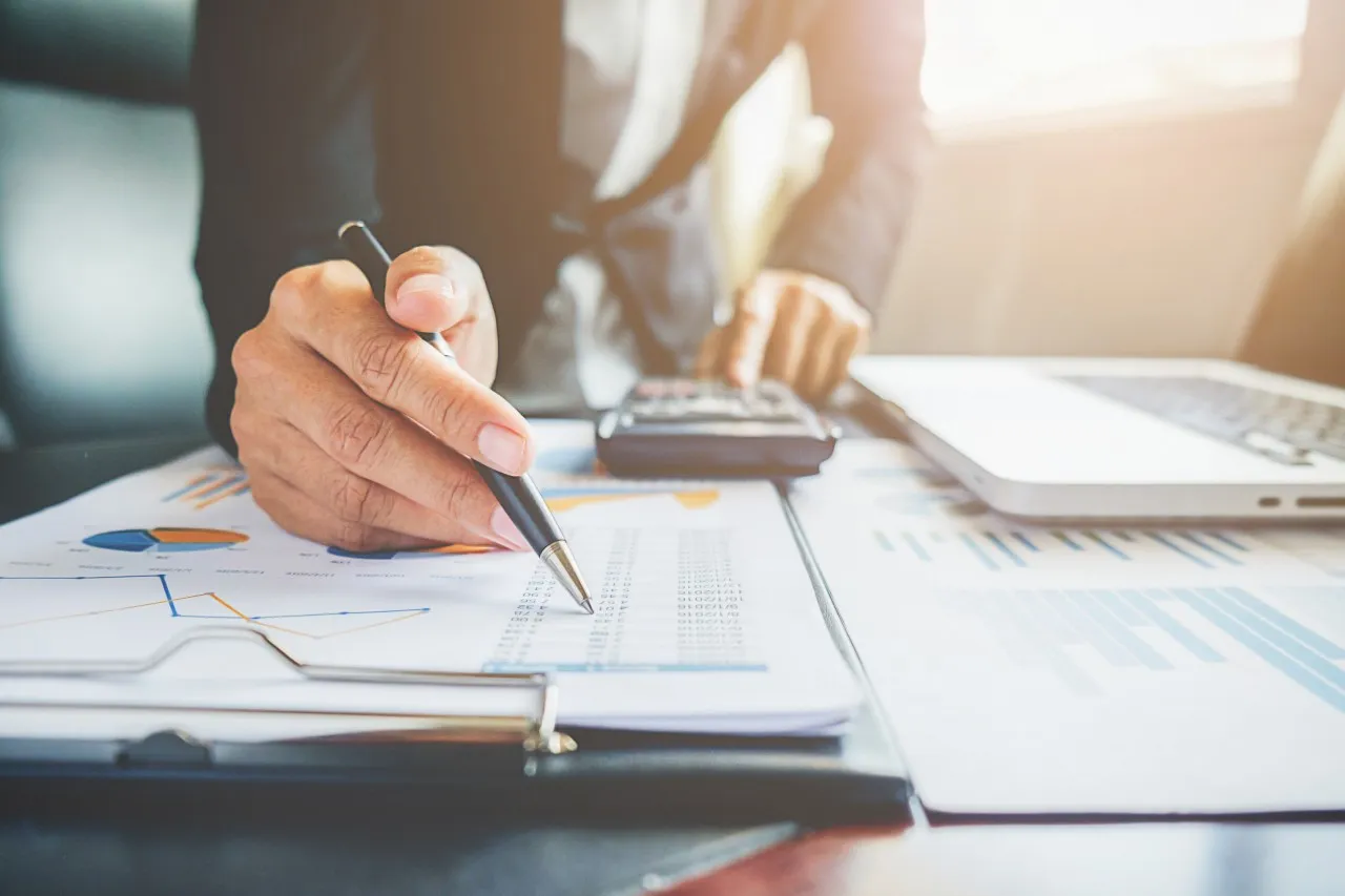 Close-up of hands holding a pen and pointing at financial documents on a clipboard. A calculator and laptop are partially visible in the background, suggesting business analysis.