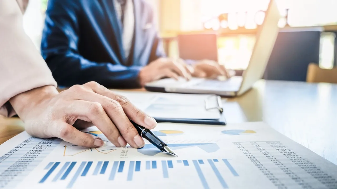 Close-up of a hand holding a pen and pointing to a bar chart on a financial document. In the blurred background, a colleague in a suit works on a laptop at a bright wooden desk.