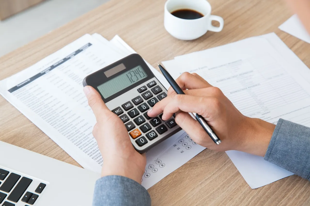 From above, hands are shown using a calculator displaying '12345' and holding a pen over paperwork on a wooden desk. A cup of coffee and part of a laptop are also visible.